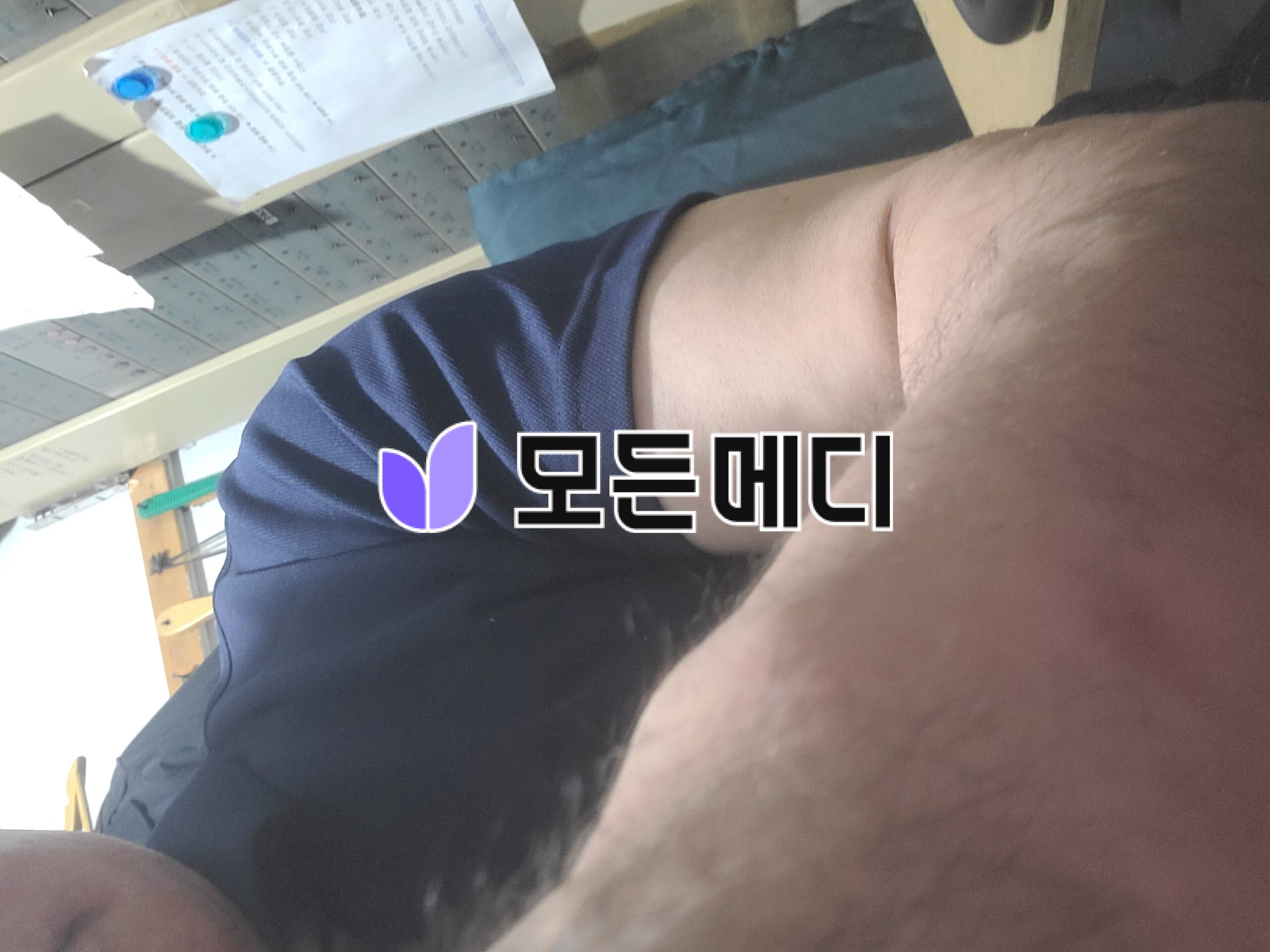 Partial view of a person wearing a navy T-shirt with a purple logo at the bottom of the frame; a desk with papers and a chair is visible on the left.