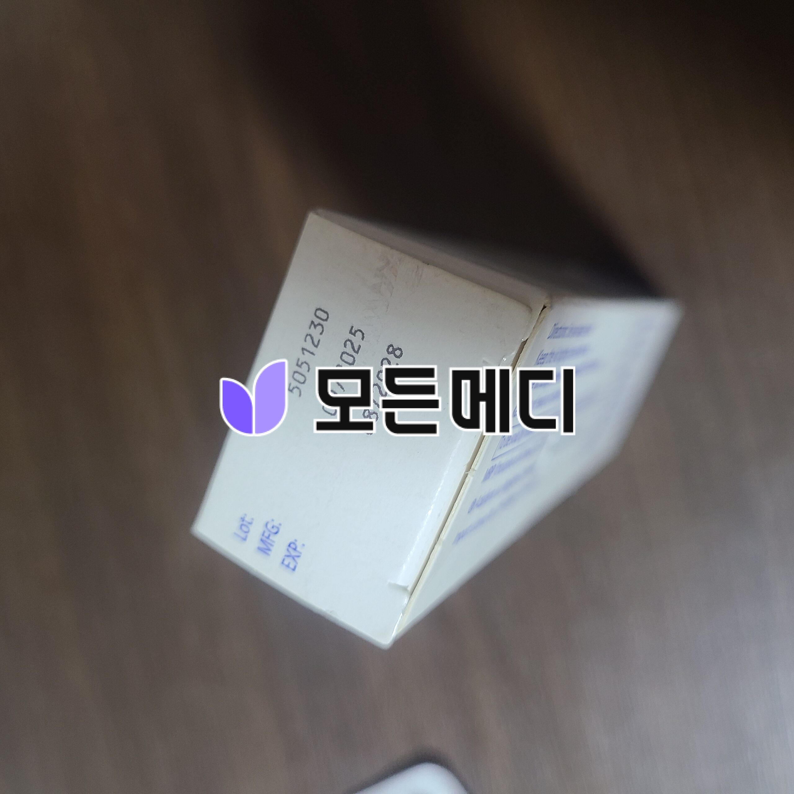 Close-up of a white box with a purple leaf-like logo on the left and bold black outlined text on a wooden surface, tilted at an angle.
